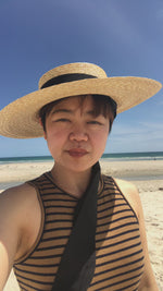 Load and play video in Gallery viewer, A woman wearing Grosgrain Amal the wide-brimmed natural straw boater hat with chin strap ribbons in the beach
